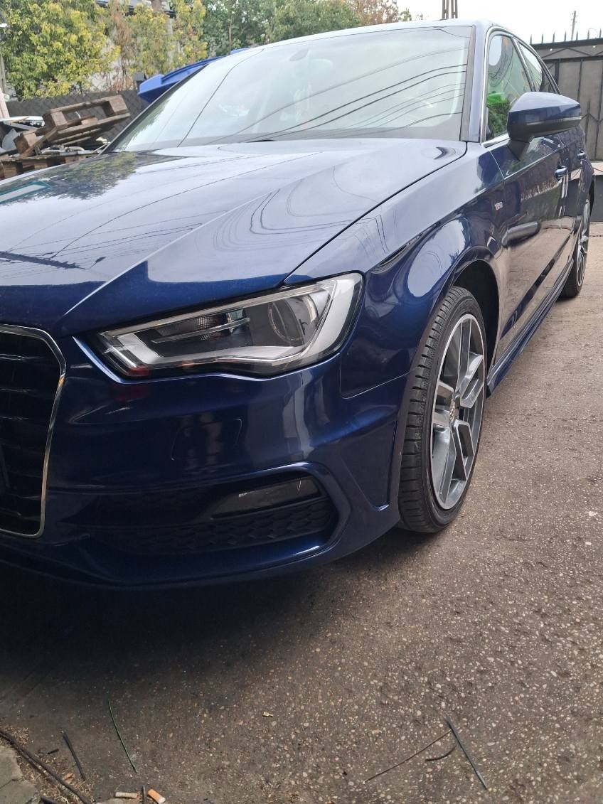 Granatowy samochód marki Audi A3, widok z przodu z lekkiego boku, na szarym betonowym podłożu. Widoczne felgi i reflektory.