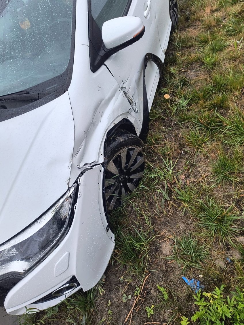 Uszkodzony przód białego samochodu marki Honda, widoczne wgniecenia i oderwany zderzak. Auto stoi na trawie, prawdopodobnie po kolizji.