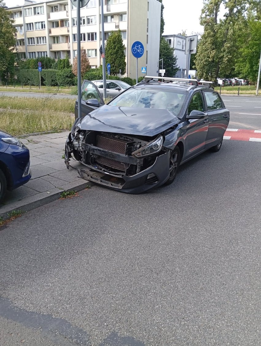 Uszkodzony, szary samochód kombi z otwartymi drzwiami po kolizji, stojący na ulicy przy chodniku. Widoczne zniszczenia przodu pojazdu i odsłonięta chłodnica.