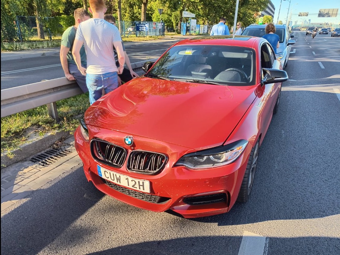 Czerwone BMW serii 2 z uszkodzonym przodem, stoi na poboczu w Rybiu. Widoczni ludzie obok pojazdu. Tablica rejestracyjna CUW 12H.