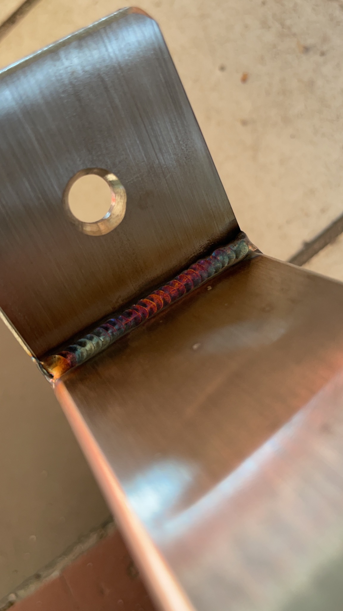 Szczegółowe zbliżenie spoiny na nierdzewnym elemencie konstrukcyjnym z widocznymi kolorami hartowania, otwór montażowy w górnym rogu.