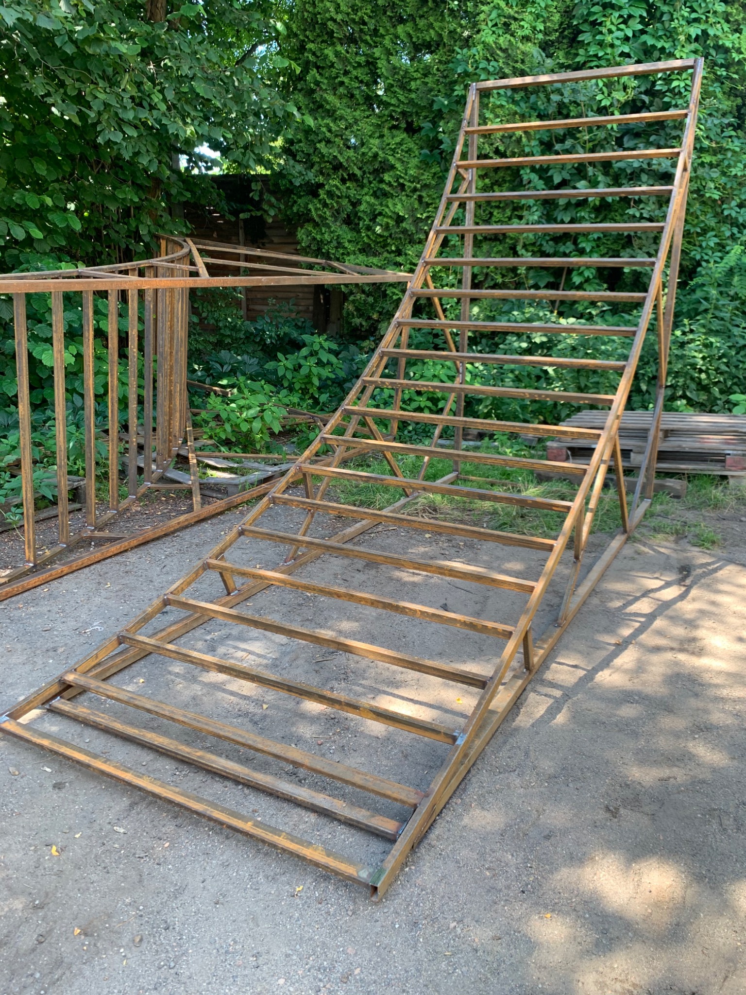 Surowa, metalowa konstrukcja schodów z elementami balustrady w tle, w trakcie budowy, z widocznymi spawami i surowym wykończeniem, ustawiona na szarym podłożu na tle zieleni.