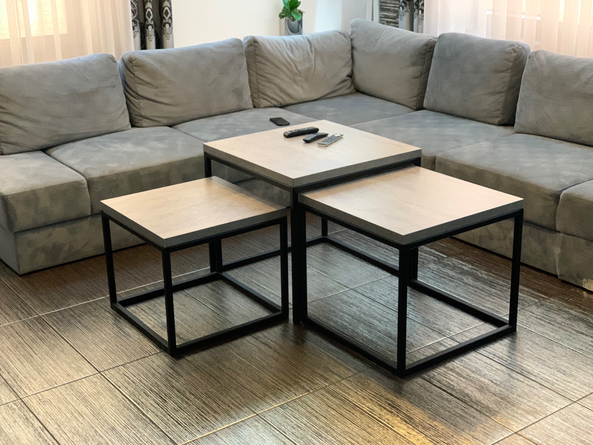 Zestaw trzech stolików kawowych o kwadratowych blatach w odcieniach beżu z czarnymi metalowymi ramami, ustawionych obok szarej, modułowej sofy w salonie z drewnopodobną podłogą z płytek.
