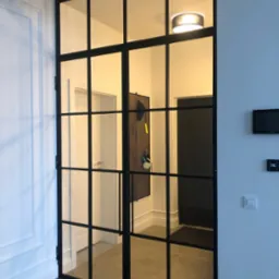 Dwuskrzydłowe, przeszklone drzwi w czarnej metalowej ramie, dzielone na prostokątne panele, oddzielające korytarz z wieszakiem na ścianie i oświetleniem sufitowym.