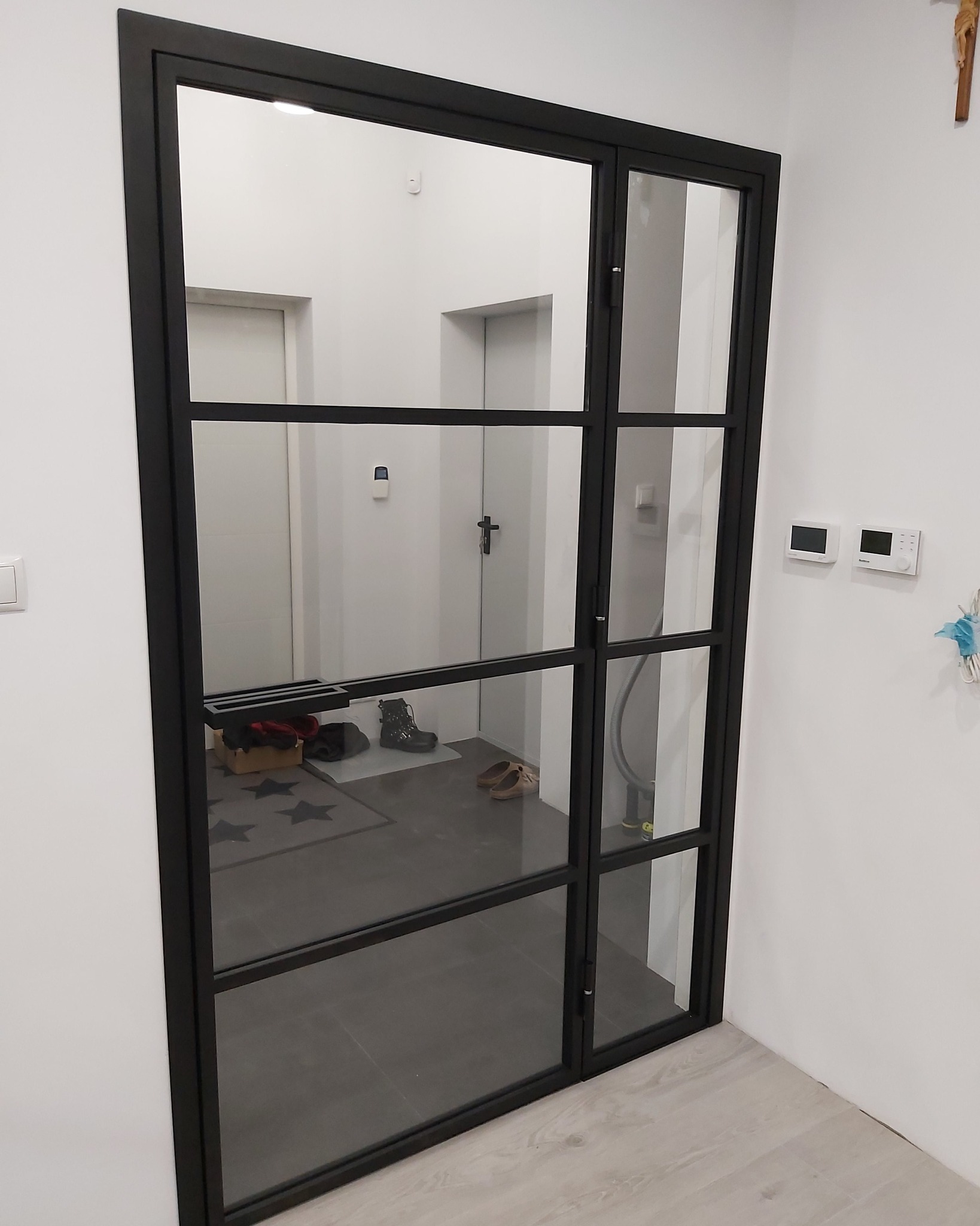 Nowoczesne, czarne, przeszklone drzwi wewnętrzne z metalową ramą, dzielące przestrzeń z widocznym przedpokojem i elementami wyposażenia.