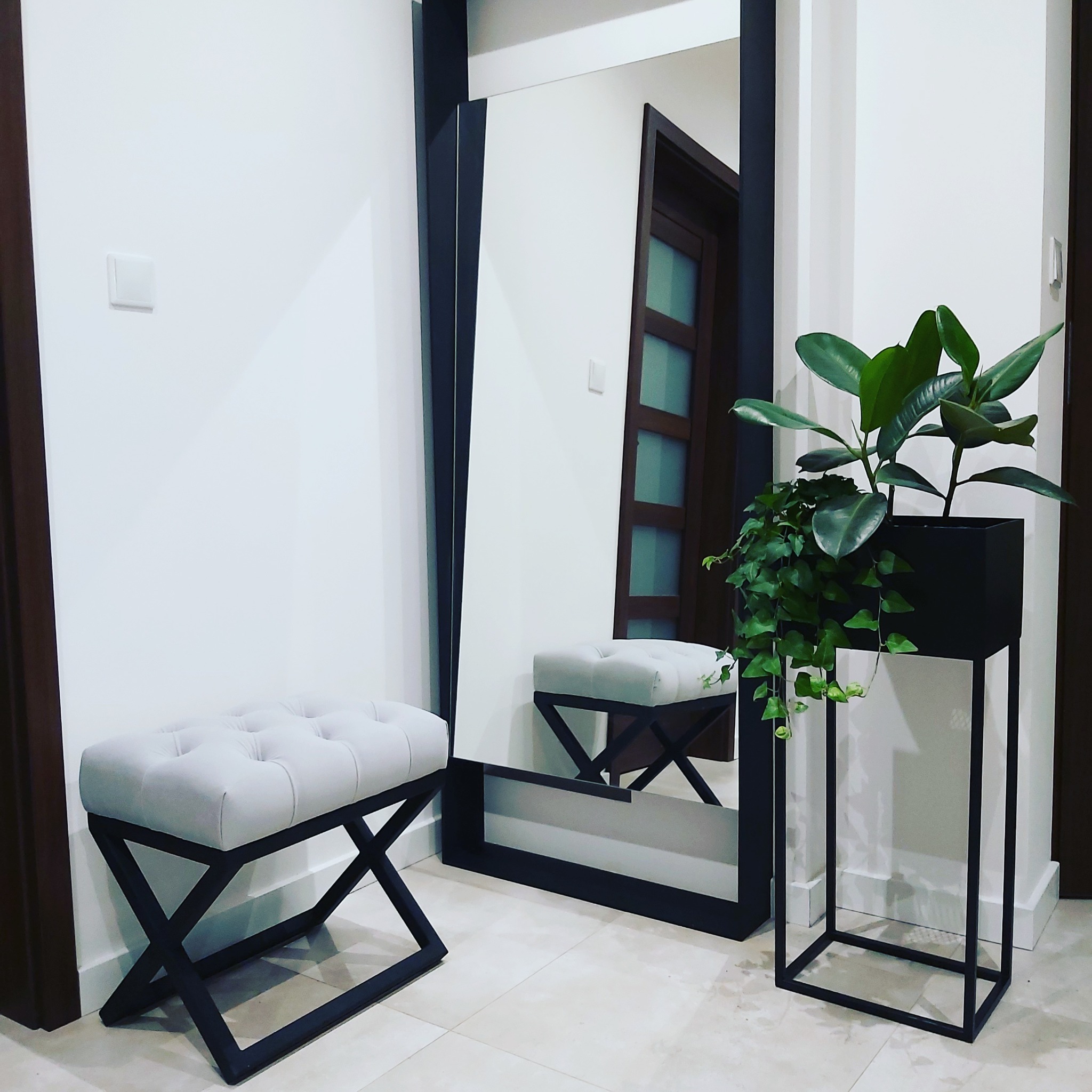 Nowoczesny przedpokój z lustrem w czarnej metalowej ramie, pufą z jasnym siedziskiem i metalową podstawą oraz stojakiem na rośliny z zielonymi liśćmi.