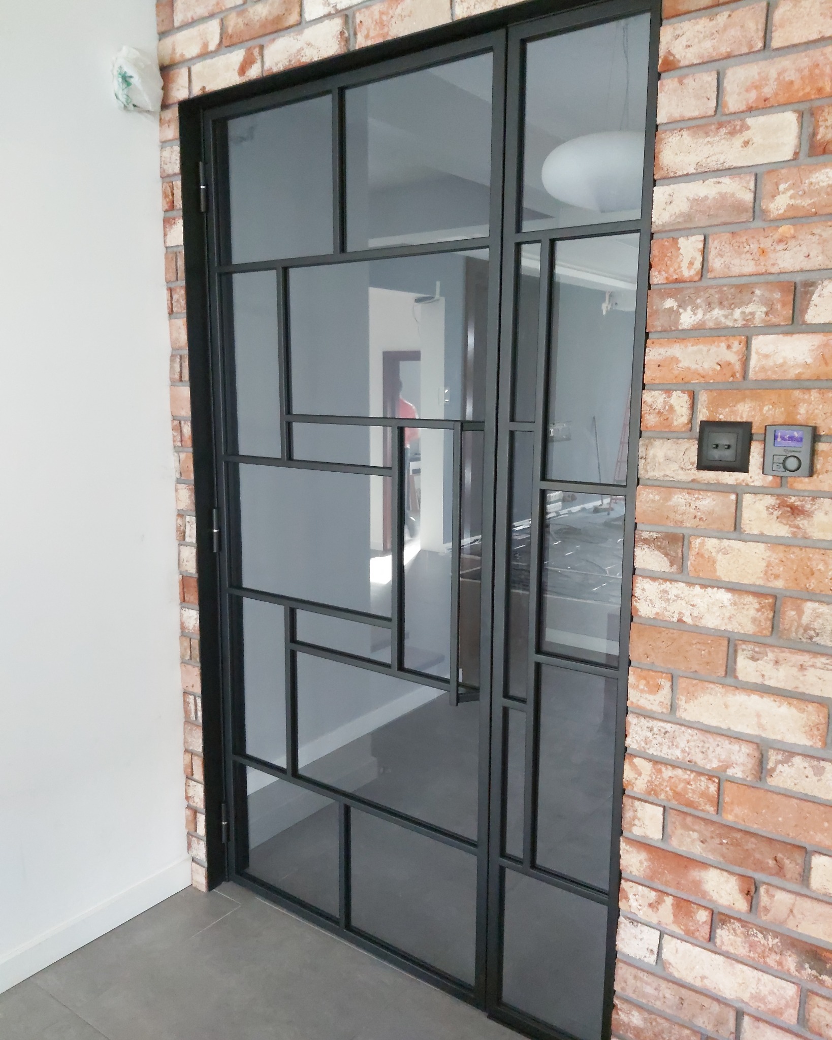 Szklane drzwi w metalowej ramie z geometrycznym podziałem szyb, osadzone w murze z czerwonej cegły, widok na wnętrze z szarą podłogą i białymi ścianami.