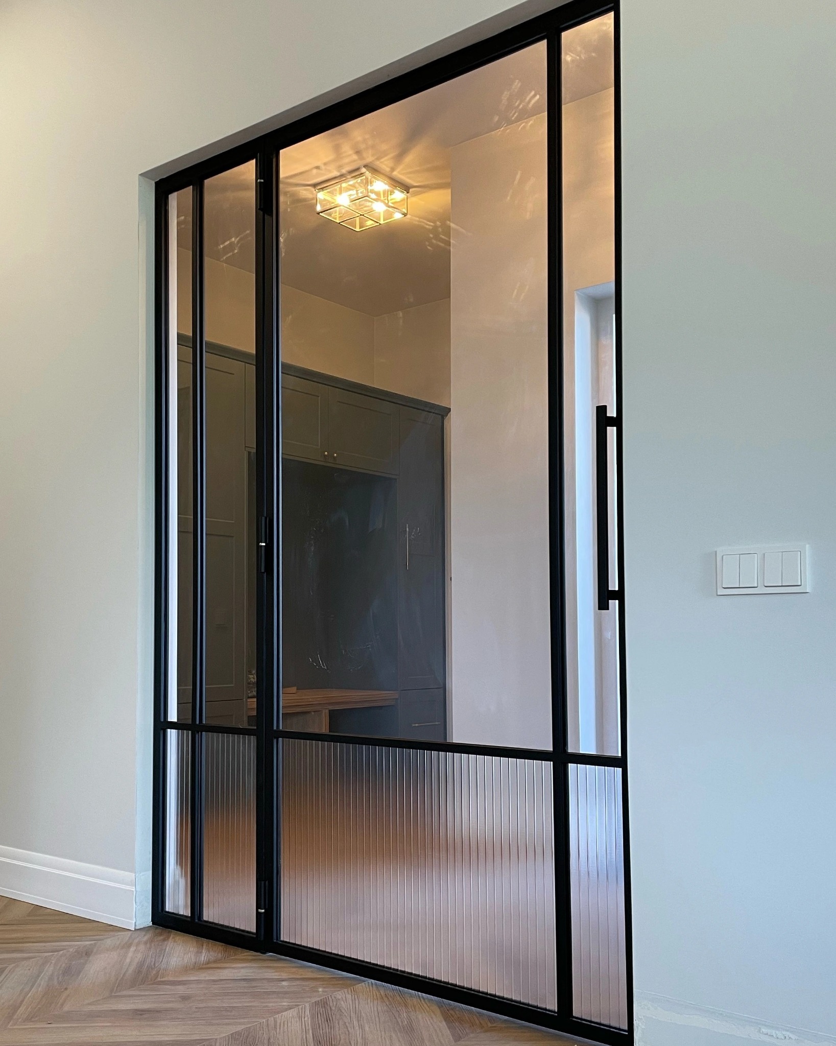 Nowoczesne drzwi wewnętrzne z czarną metalową ramą i szkłem ryflowanym, widoczne elementy wnętrza w tle, parkiet ułożony w jodełkę.