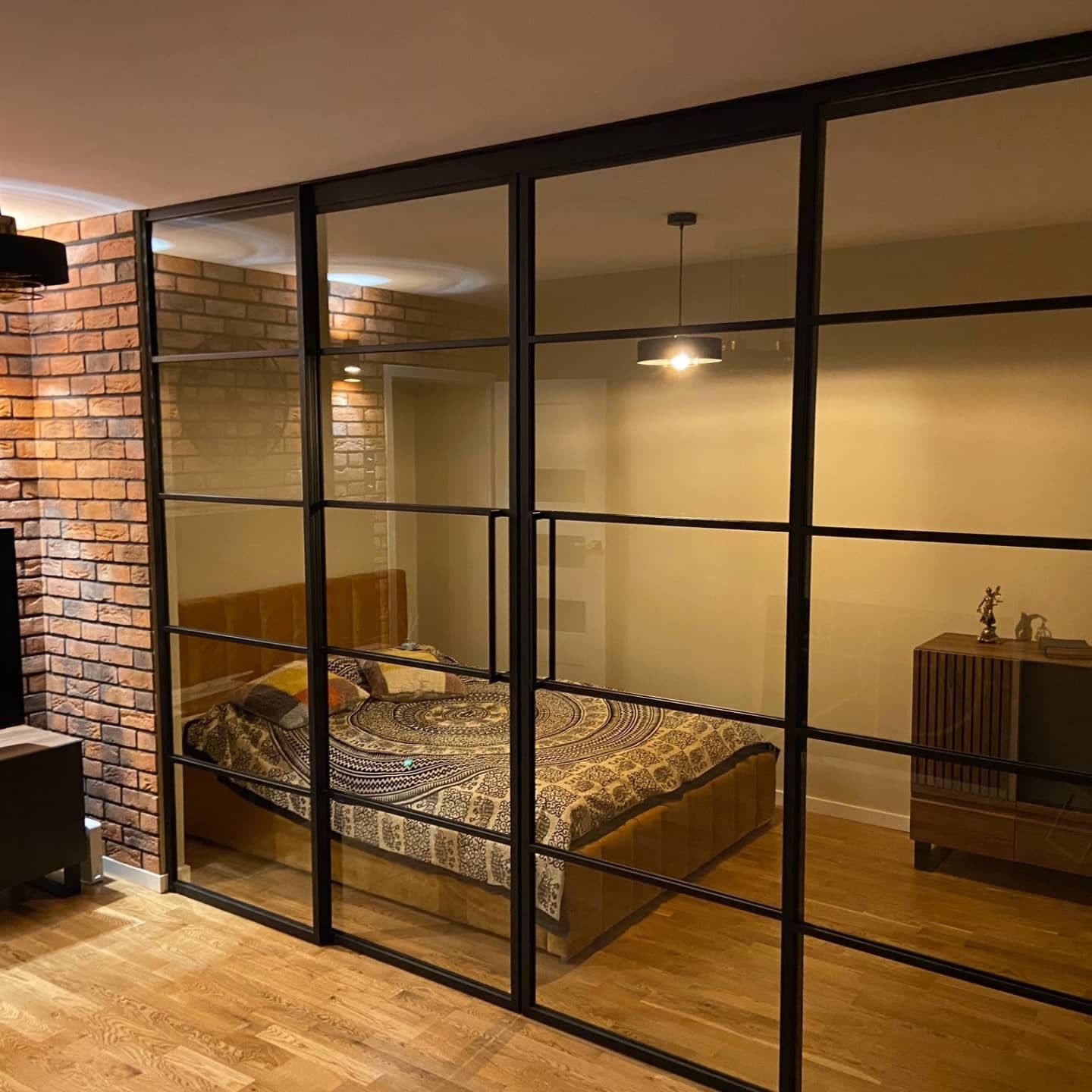 Przeszklona ściana działowa z czarnymi metalowymi profilami oddzielająca sypialnię od reszty mieszkania, widoczna ceglana ściana i drewniana podłoga.