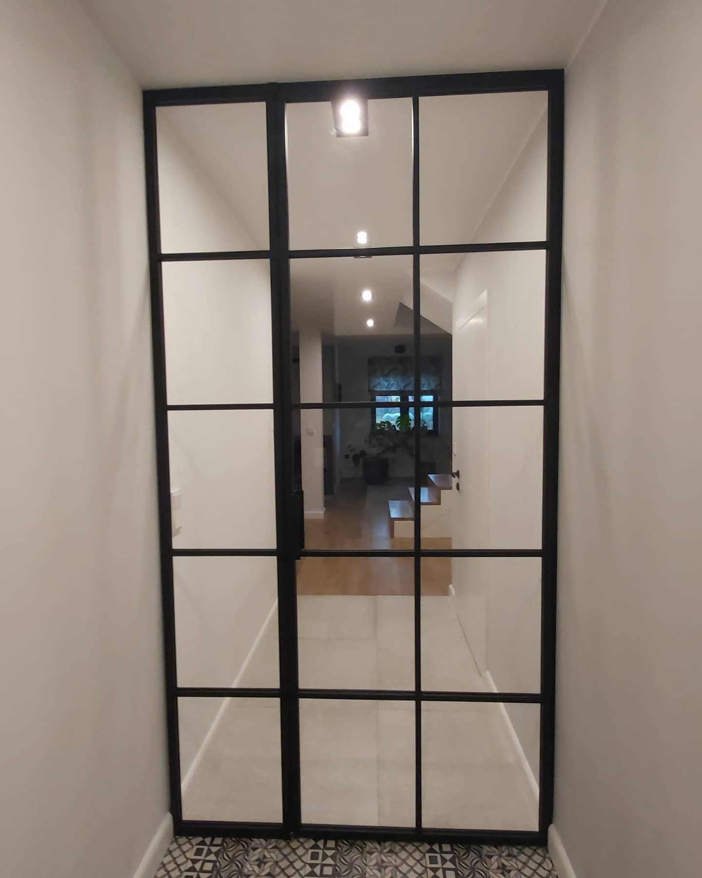 Czarna, przeszklona ścianka działowa z metalową ramą oddzielająca korytarz z wzorzystą podłogą od dalszej części domu z drewnianymi schodami i oświetleniem punktowym.