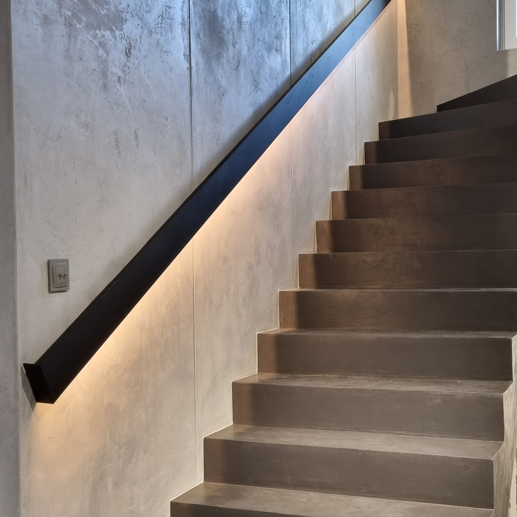 Nowoczesne schody z betonu architektonicznego z czarną, metalową poręczą podświetloną od spodu ciepłym światłem. Widoczny fragment ściany z efektem betonu oraz włącznik światła.