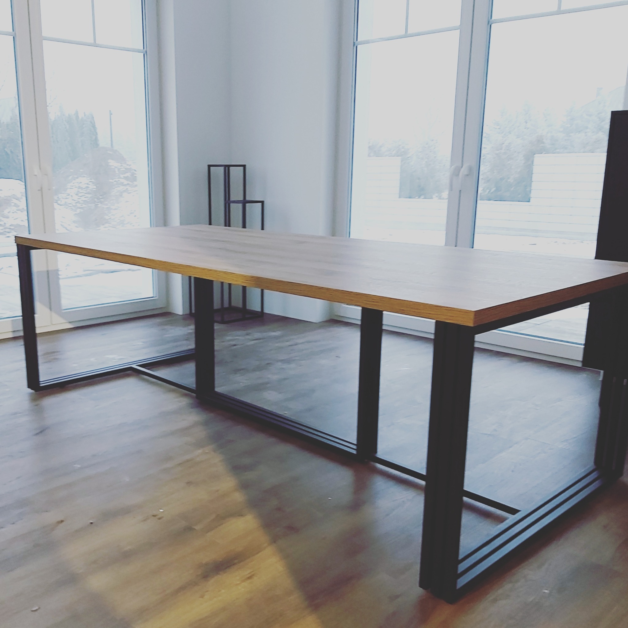 Stół z drewnianym blatem i czarną, metalową konstrukcją nóg, ustawiony w jasnym pomieszczeniu z dużymi oknami.