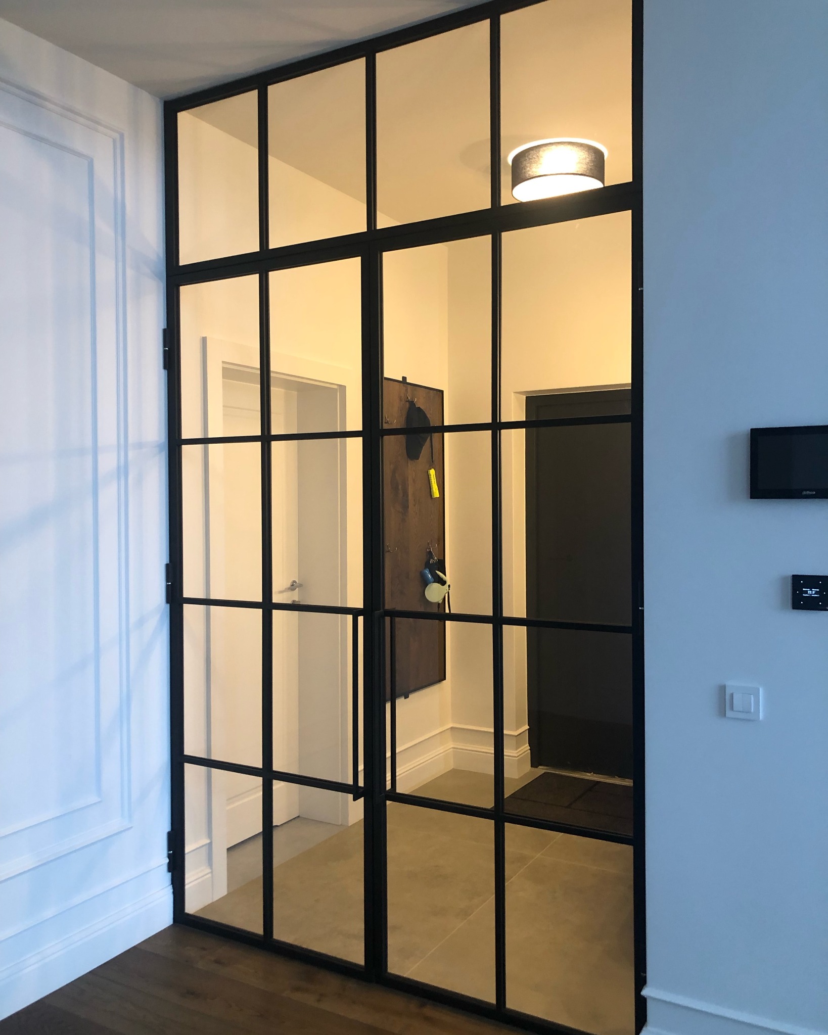 Dwuskrzydłowe, przeszklone drzwi w czarnej metalowej ramie, dzielone na prostokątne panele, oddzielające korytarz z wieszakiem na ścianie i oświetleniem sufitowym.