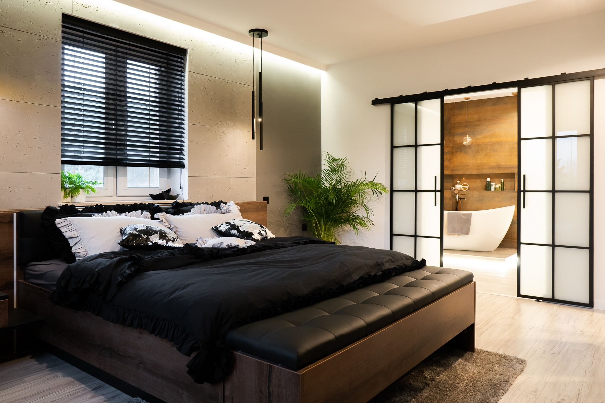 Nowoczesna sypialnia z łóżkiem z ciemnego drewna, czarną pościelą z falbanami i dekoracyjnymi poduszkami, widok na łazienkę przez przesuwne drzwi z metalową ramą w stylu industrialnym.