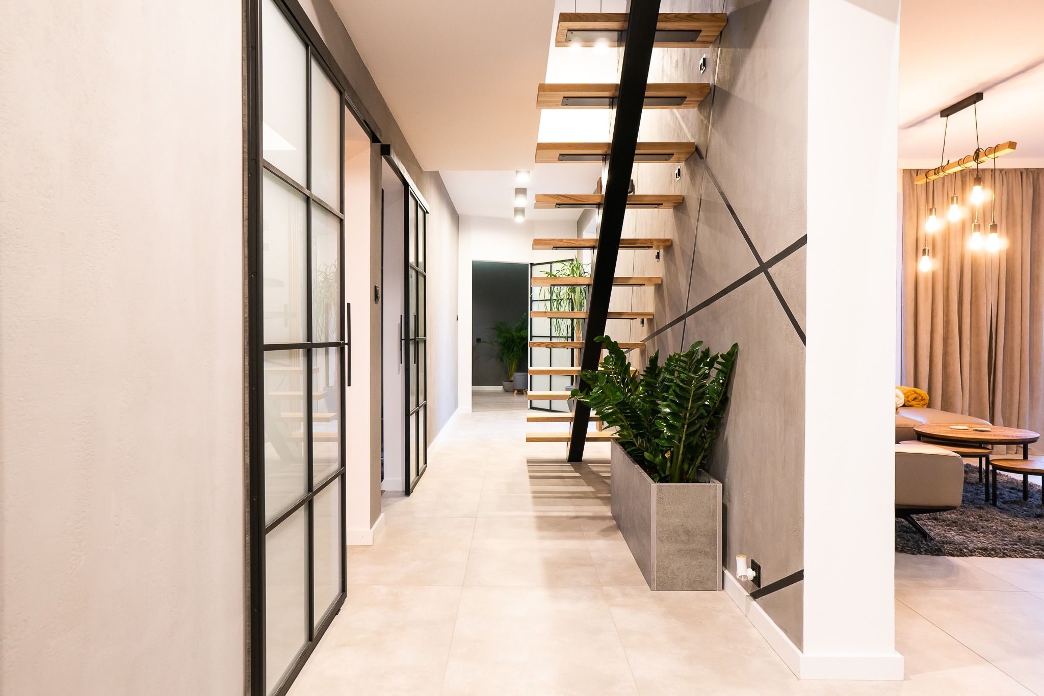 Nowoczesne schody z drewnianymi stopniami i czarną metalową konstrukcją, minimalistyczne wnętrze z przeszklonymi drzwiami i roślinami doniczkowymi.
