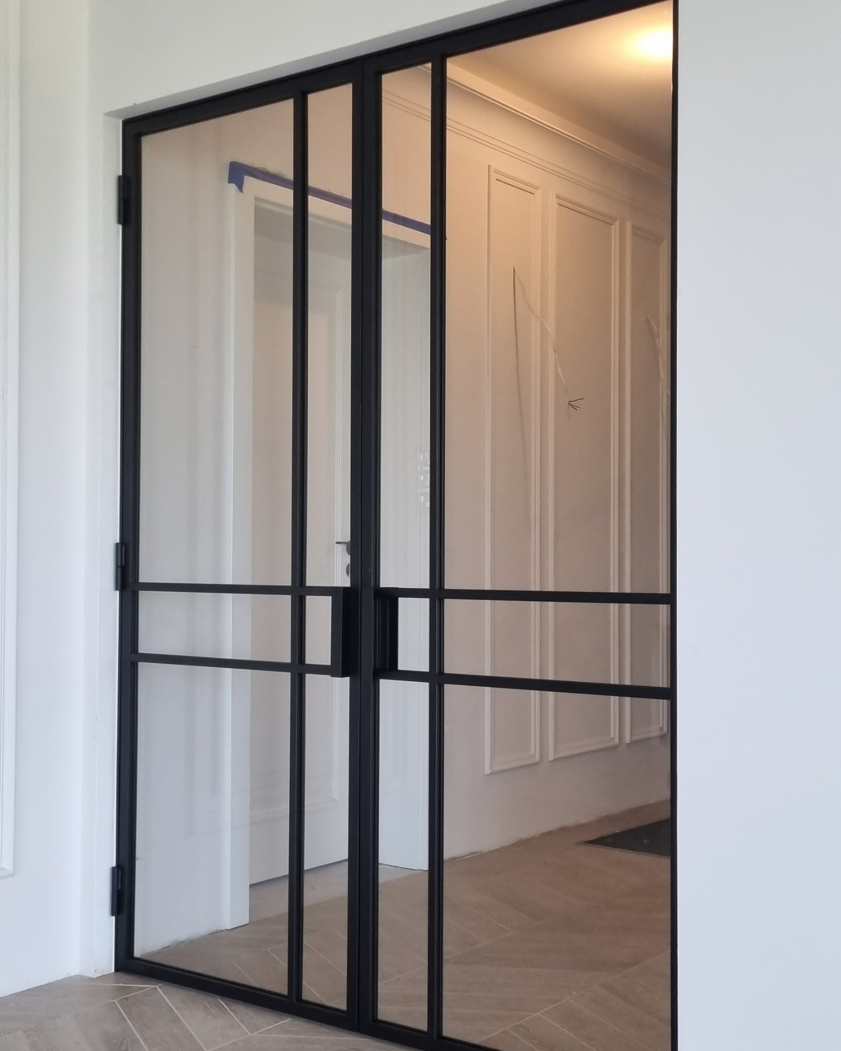 Dwuskrzydłowe drzwi z czarną, metalową ramą i szklanymi panelami, widoczne z bliska, z taśmą malarską na górnej krawędzi ościeżnicy po lewej stronie.