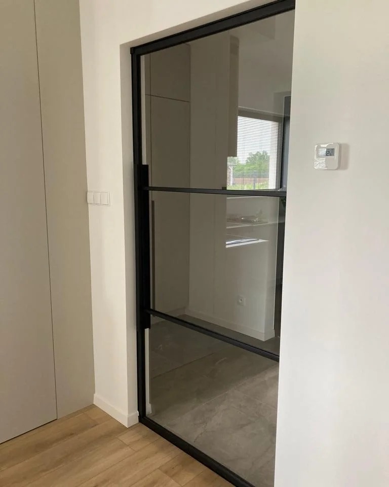 Nowoczesne drzwi wewnętrzne z czarną, metalową ramą i szklanym wypełnieniem, dzielone poziomymi szprosami, widoczne na tle jasnego wnętrza z marmurową podłogą.