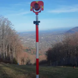 Tyczka geodezyjna z pryzmatem na wzgórzu, w tle panorama miasta i błękitne niebo.