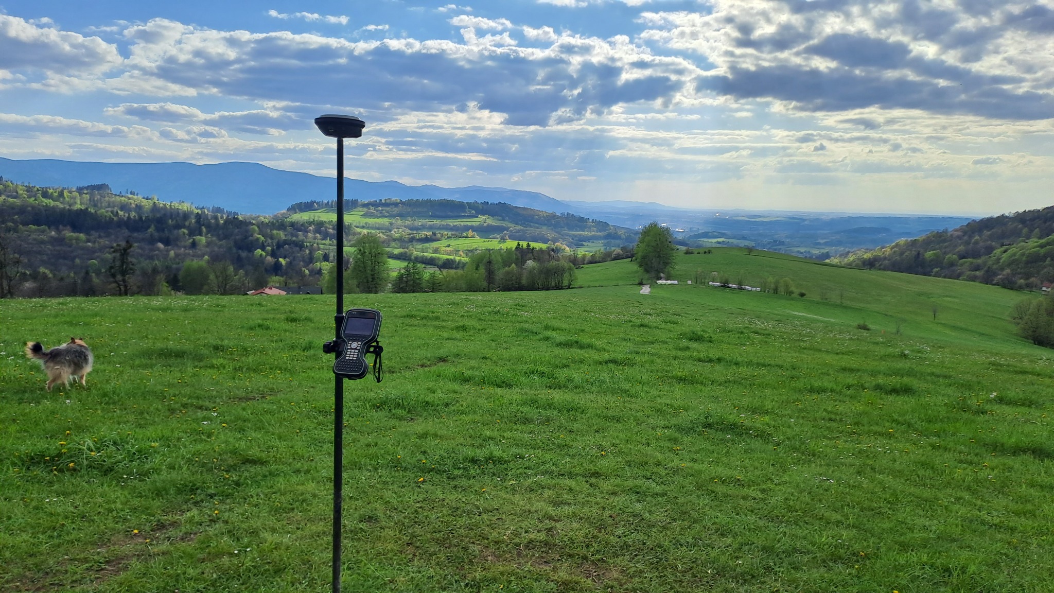 Terenowy odbiornik GNSS na tyczce z kontrolerem w uchwycie, na tle zielonej łąki z widokiem na zalesione wzgórza i błękitne niebo z chmurami. W lewym dolnym rogu widoczny pies.