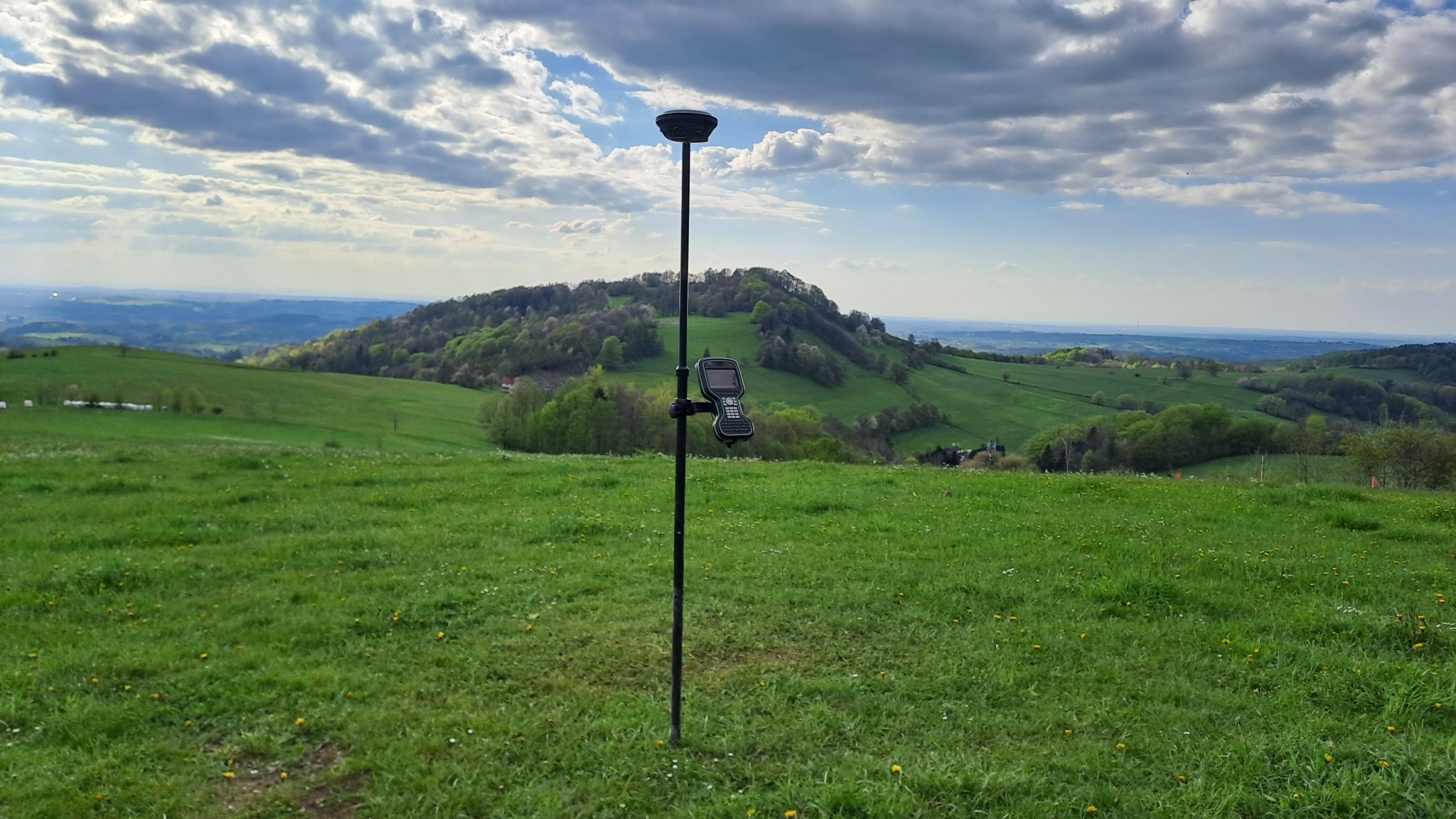 Czarny statyw z odbiornikiem GPS i kontrolerem na tle zielonego pagórkowatego krajobrazu pod częściowo pochmurnym niebem.