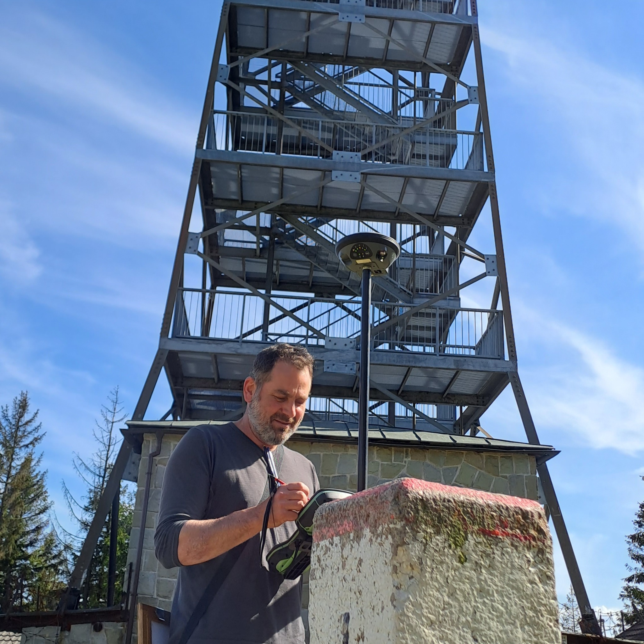 Mężczyzna z brodą, w szarej koszulce, notuje coś na urządzeniu geodezyjnym stojącym na betonowym słupku z czerwoną linią, na tle stalowej wieży widokowej i błękitnego nieba.