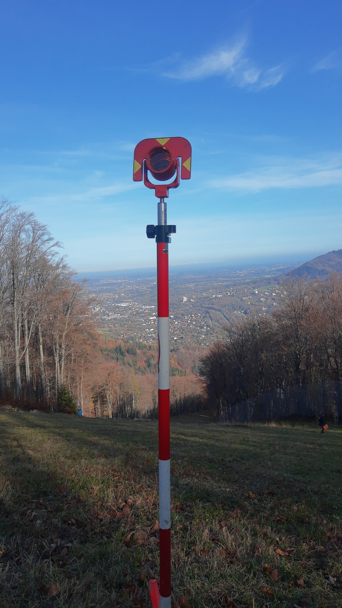 Tyczka geodezyjna z pryzmatem na wzgórzu, w tle panorama miasta i błękitne niebo.