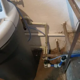 Szara stacja uzdatniania wody z podłączonymi rurami z tworzywa sztucznego w piwnicy, widoczne złącza i zawory, w tle ściana z odpadającym tynkiem.