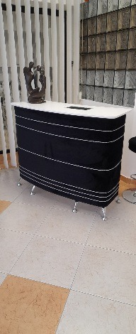 Recepcja w kolorze czarnym z białym blatem i srebrnymi nogami, na blacie rzeźba przedstawiająca parę, w tle ściana z dekoracyjnych płytek.