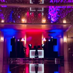 Scena z nagłośnieniem i oświetleniem w fioletowo-niebieskich barwach, lustrzana kula pod sufitem, napis 'Let's Dance!' na podeście DJ-a.