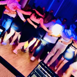 Tańczący w kręgu ludzie, oświetleni niebieskim światłem, z widocznym laptopem DJ-a w rogu kadru. Parkiet, impreza, zabawa.