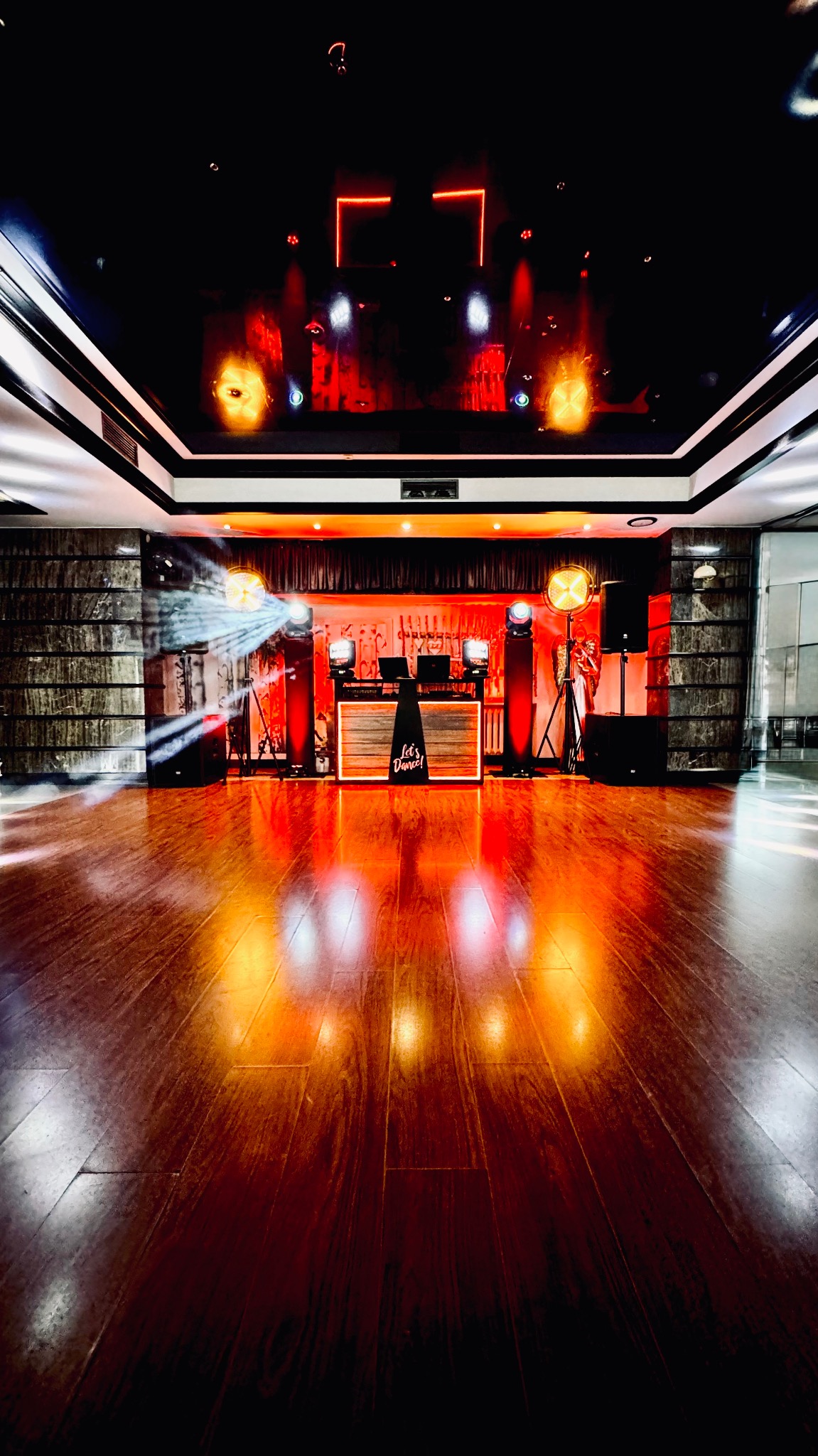 Sala z parkietem, oświetleniem scenicznym i stanowiskiem DJ-a z napisem 'Let's Dance!', gotowa na imprezę. Czerwone światła odbijają się w podłodze.