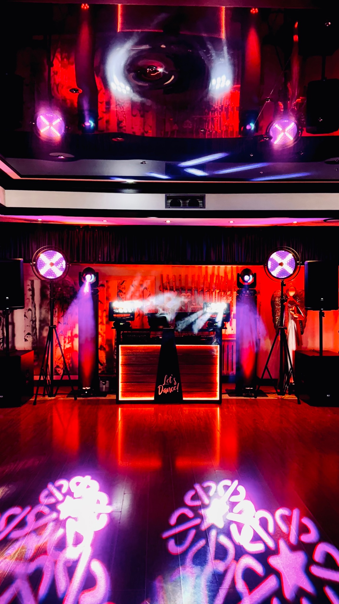 Scena oświetlona czerwonym światłem z dekoracjami świetlnymi na parkiecie, sprzętem DJ-skim i reflektorami na statywach. Na scenie napis 'Let's Dance!'.