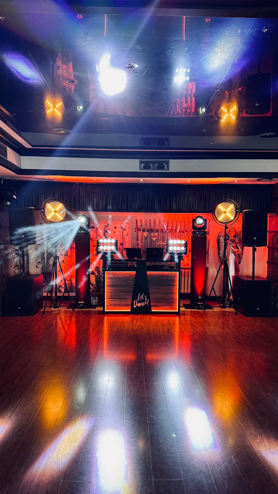 Scena z konsoletą DJ-ską, oświetleniem scenicznym i nagłośnieniem w lokalu. Na parkiecie widoczne refleksy świetlne. Na blacie konsolety widoczny laptop.
