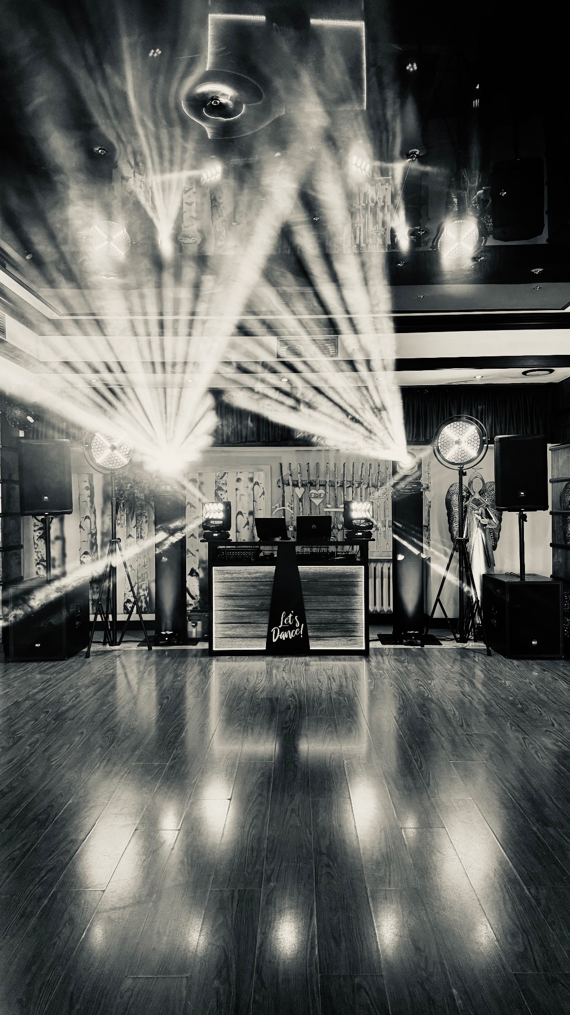 Czarno-białe zdjęcie: stanowisko DJ'a z napisem 'Let's Dance!', głośniki, reflektory i efekty świetlne na parkiecie. Widoczne elementy dekoracyjne i oświetlenie sufitowe.