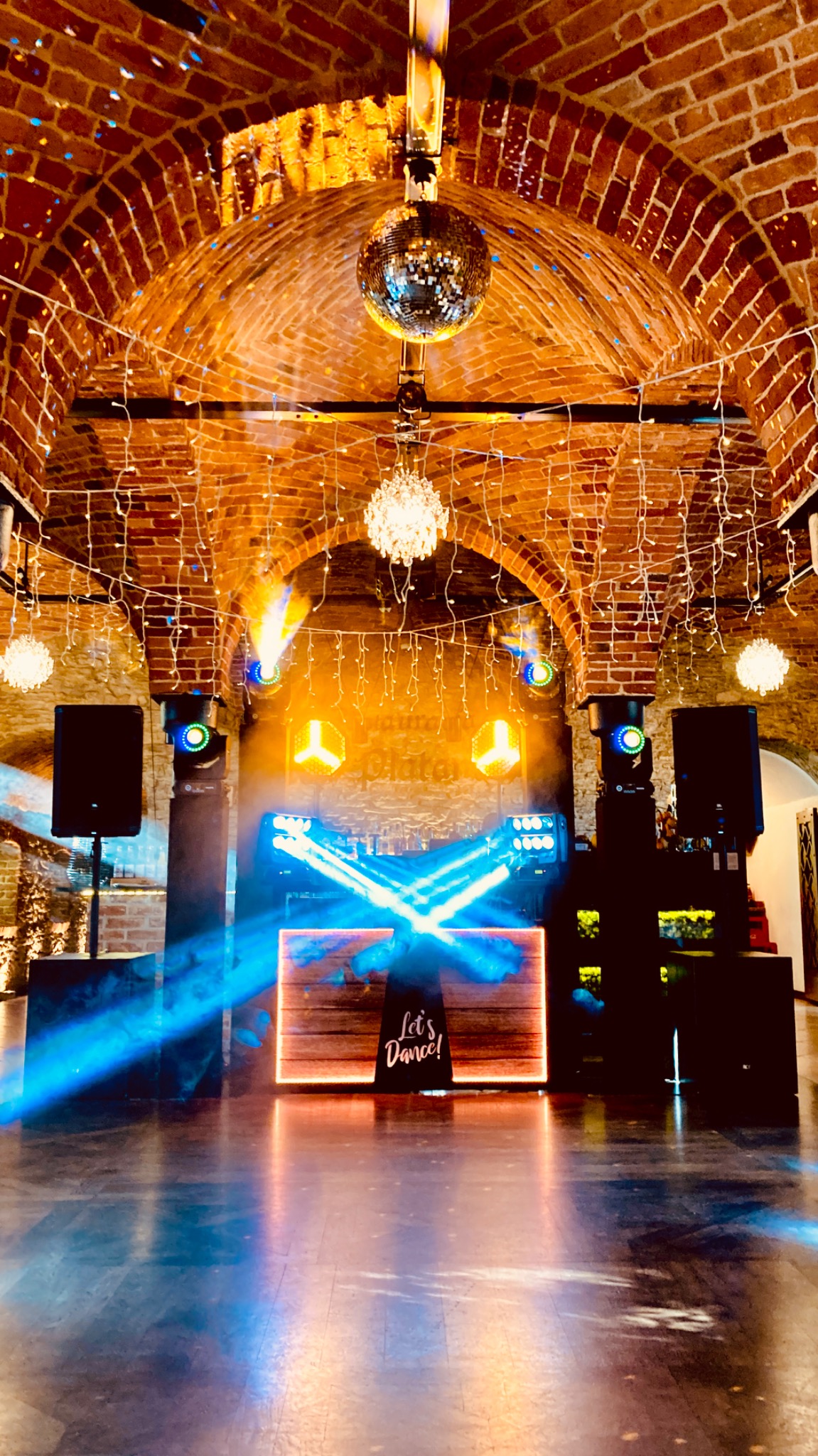 Oświetlenie eventowe w ceglanym wnętrzu z parkietem: reflektory, kule dyskotekowe i dekoracyjne światełka tworzą klimat imprezy. DJ'ka z napisem 'Let's Dance!'.