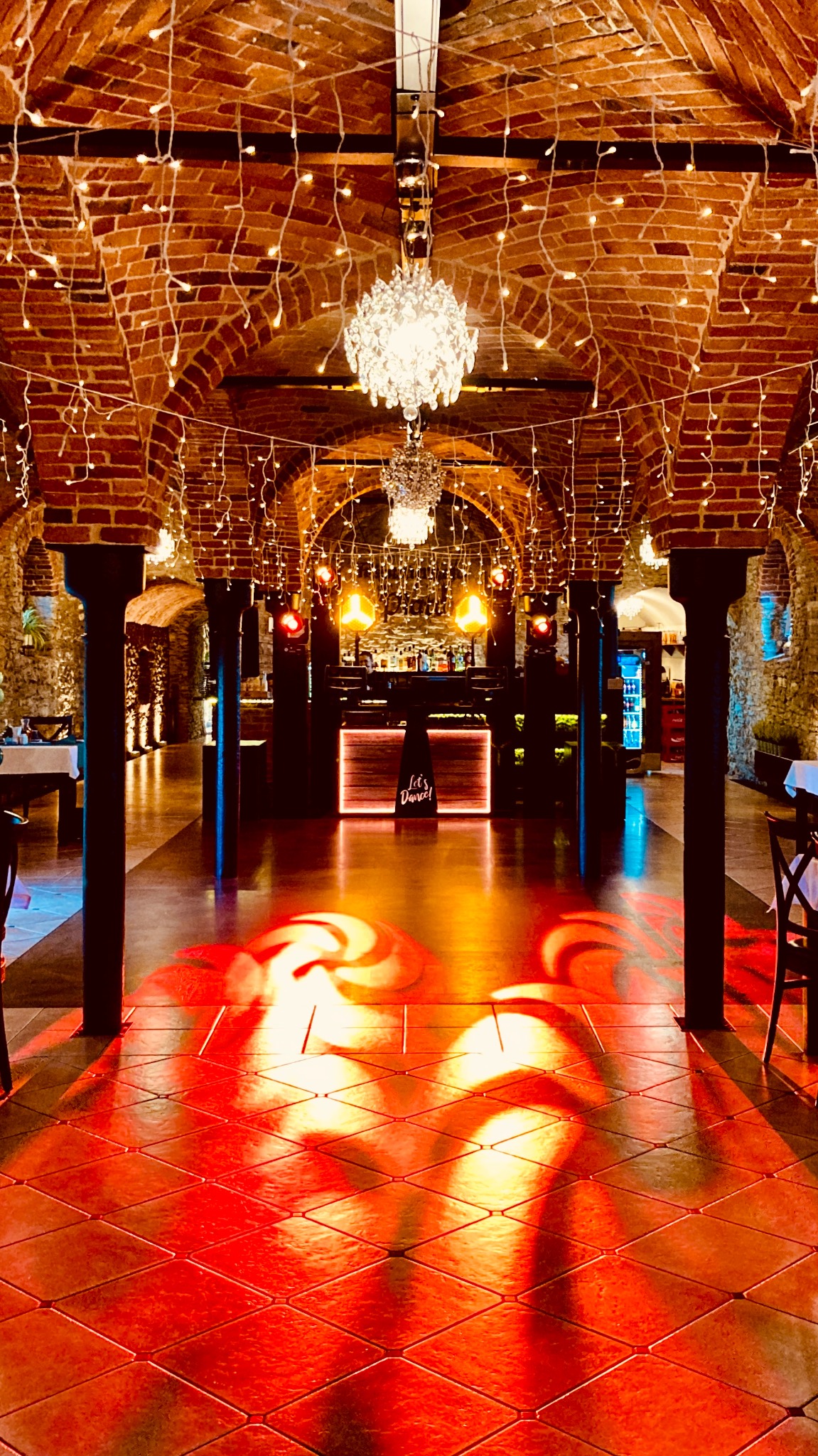 Sala balowa z ceglanymi łukami, oświetlona girlandami i żyrandolami, parkiet w czerwonej poświacie, bar w tle. Gotowa przestrzeń na imprezę.