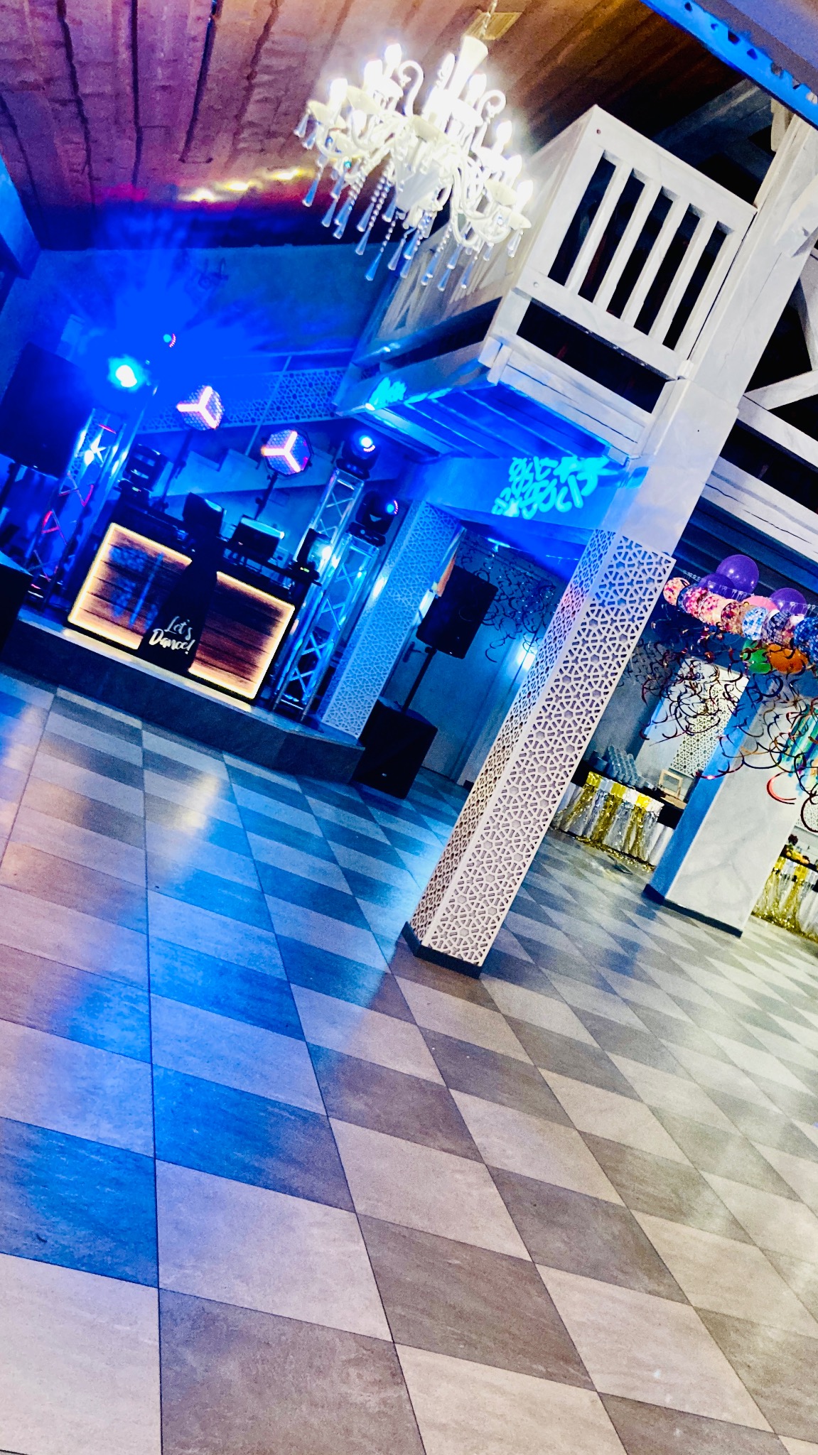 Sala bankietowa z parkietem w szachownicę, oświetlona niebieskim światłem. Widoczny DJ na podeście, dekoracje i balony. Gotowa na imprezę.