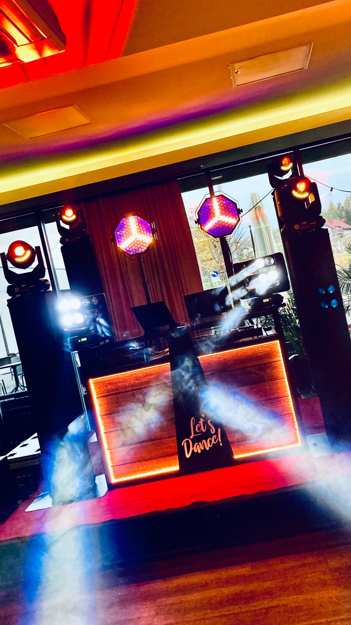 Scena z DJ-em, oświetleniem i napisem 'Let's Dance!'. Kolorowe światła tworzą dynamiczną atmosferę imprezową. Widoczne elementy nagłośnienia i efekty dymne.