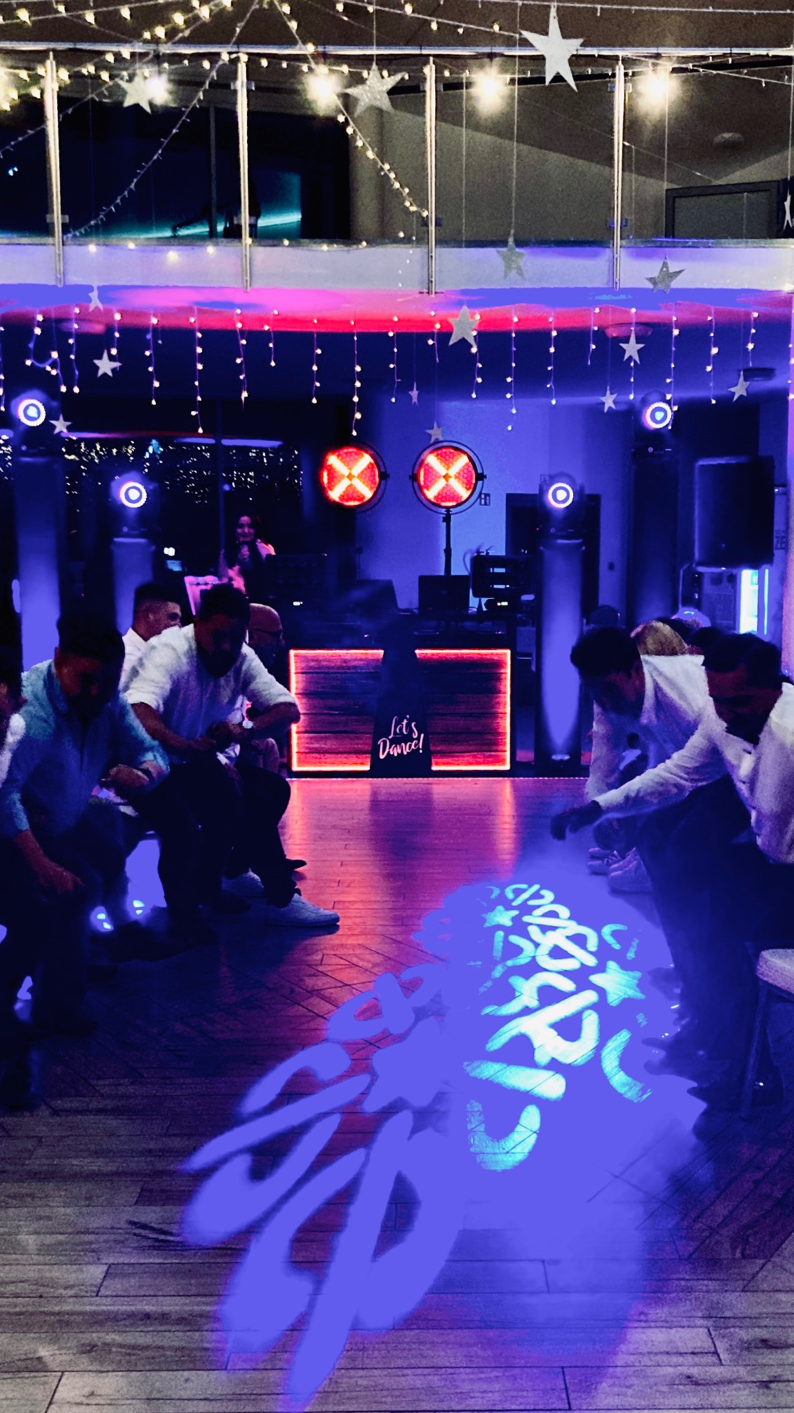 Parkiet taneczny oświetlony niebieskimi wzorami, w tle DJ-ka z napisem 'Let's Dance!', dekoracje świetlne i bawiący się ludzie. Scena z imprezy.