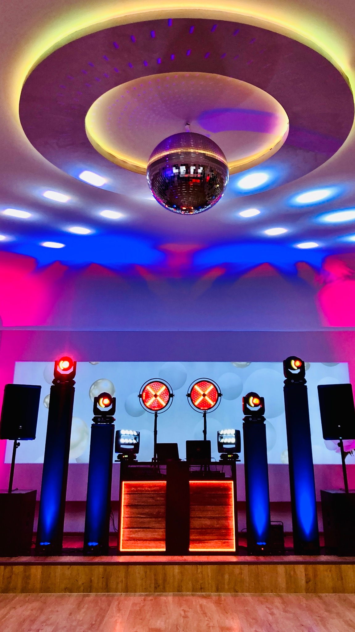 Scena z nagłośnieniem i oświetleniem: kolumna głośnikowa, reflektory sceniczne, konsoleta DJ-ska. W tle ściana z dekoracjami i kula dyskotekowa pod sufitem.