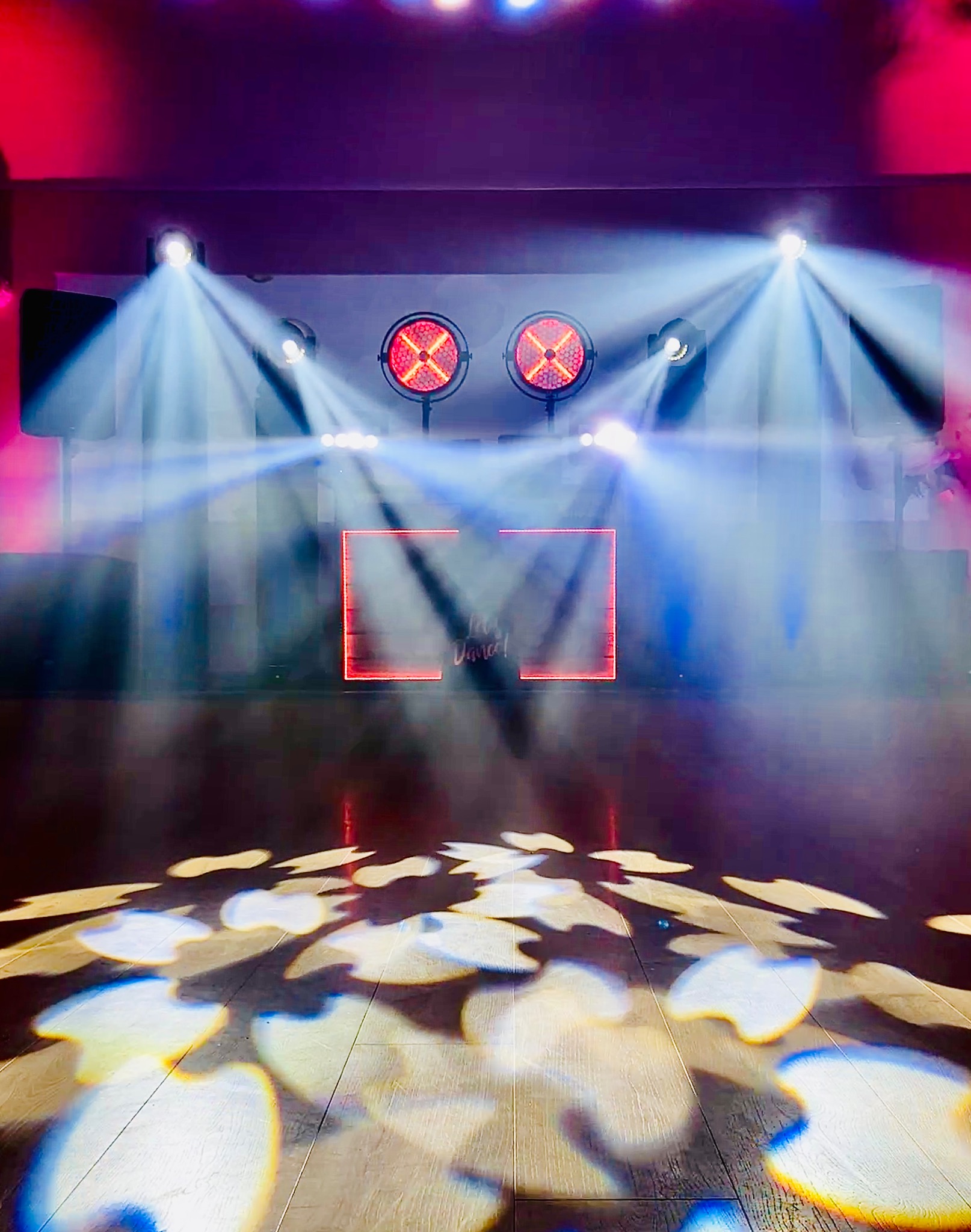 Scena oświetlona reflektorami punktowymi i dwoma okrągłymi lampami z czerwonymi krzyżami, z napisem 'Let's Dance!' w neonowej ramce.