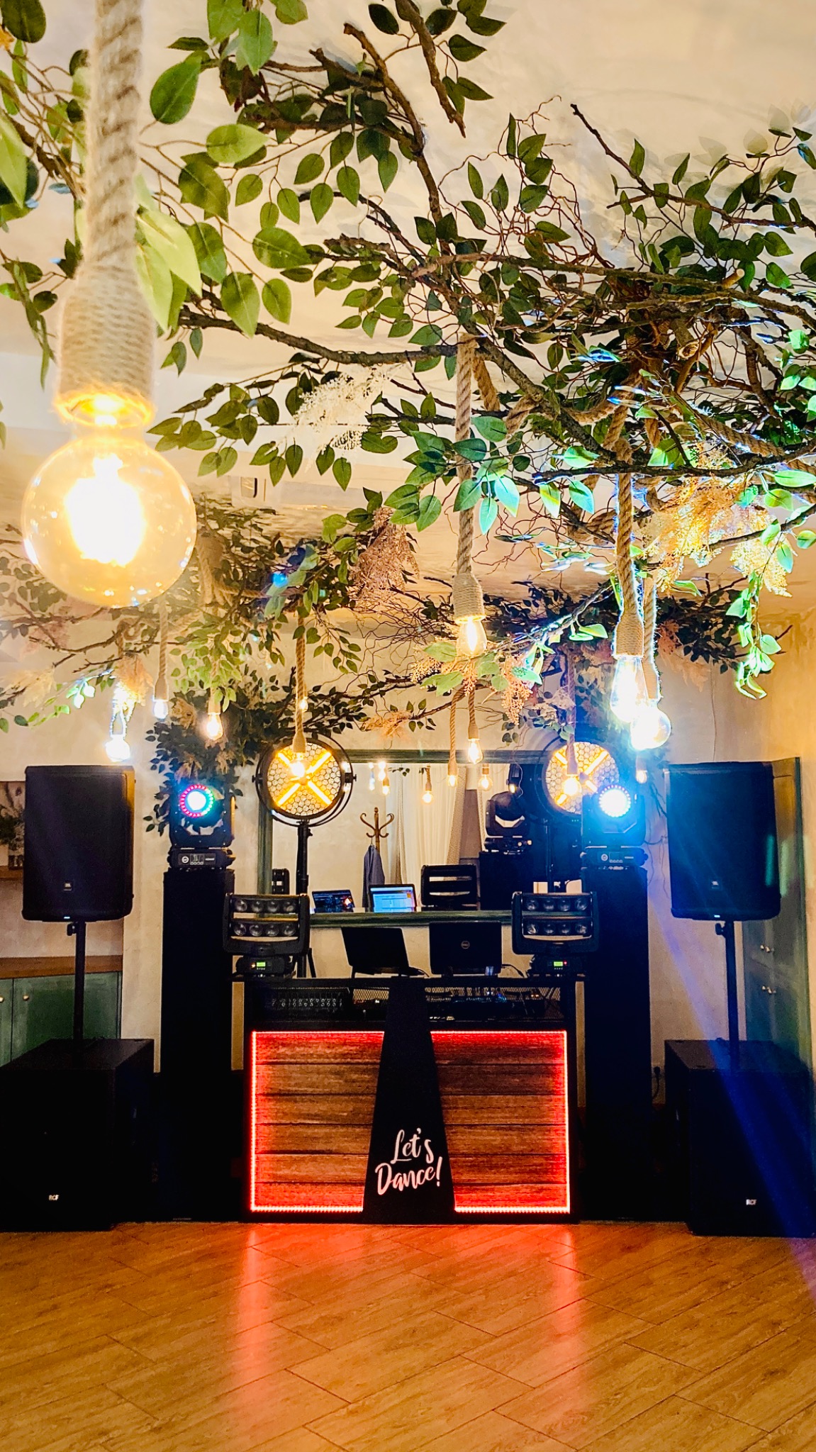 Dekoracja sali z nagłośnieniem i oświetleniem eventowym: kolumny głośnikowe, konsoleta DJ-ska z napisem 'Let's Dance!', dekoracje roślinne i wiszące żarówki.
