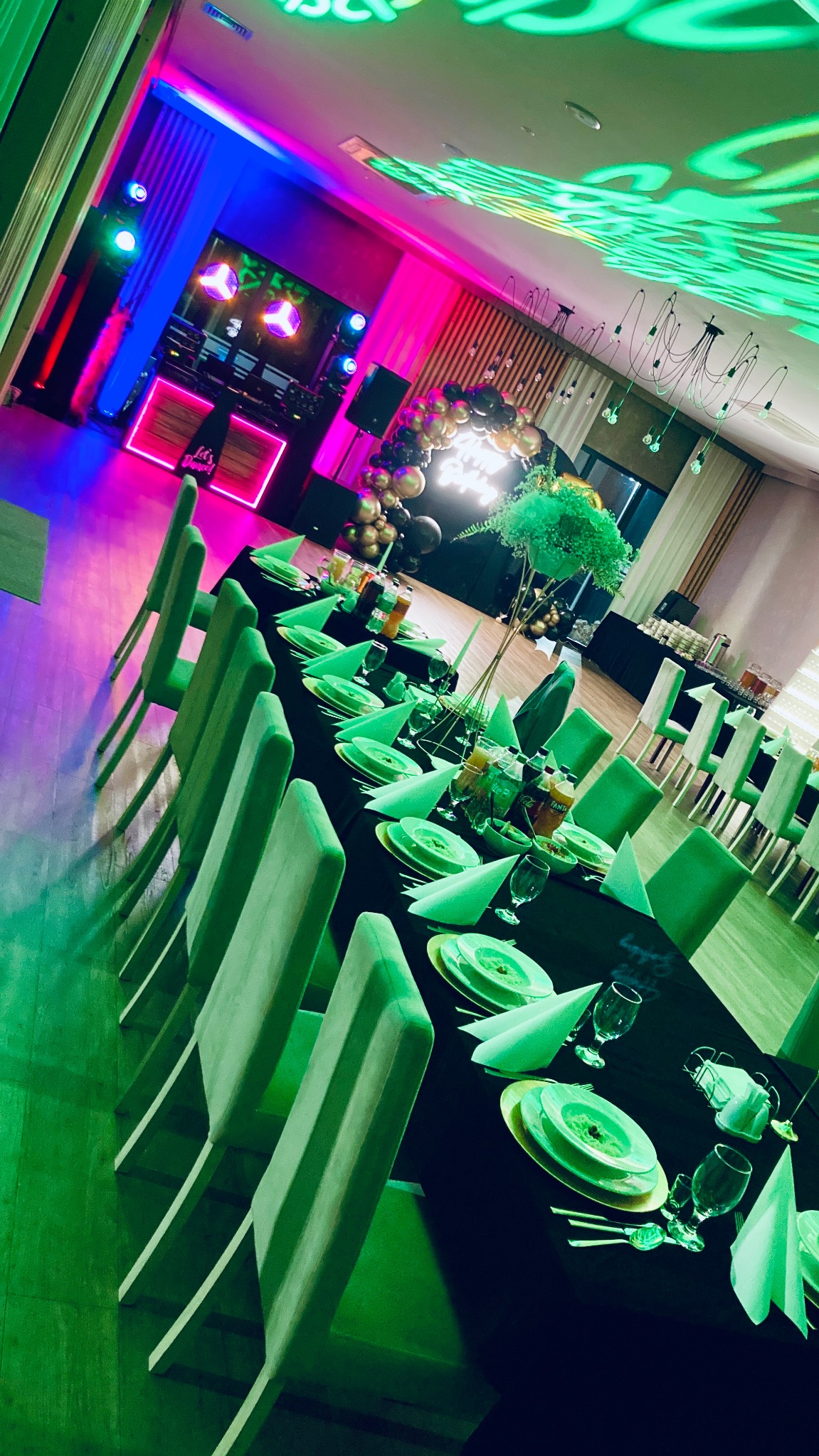 Uroczysta sala bankietowa z długim stołem, nakrytym zieloną zastawą i czarnym obrusem, oświetlona kolorowymi światłami, z dekoracjami balonowymi w tle.