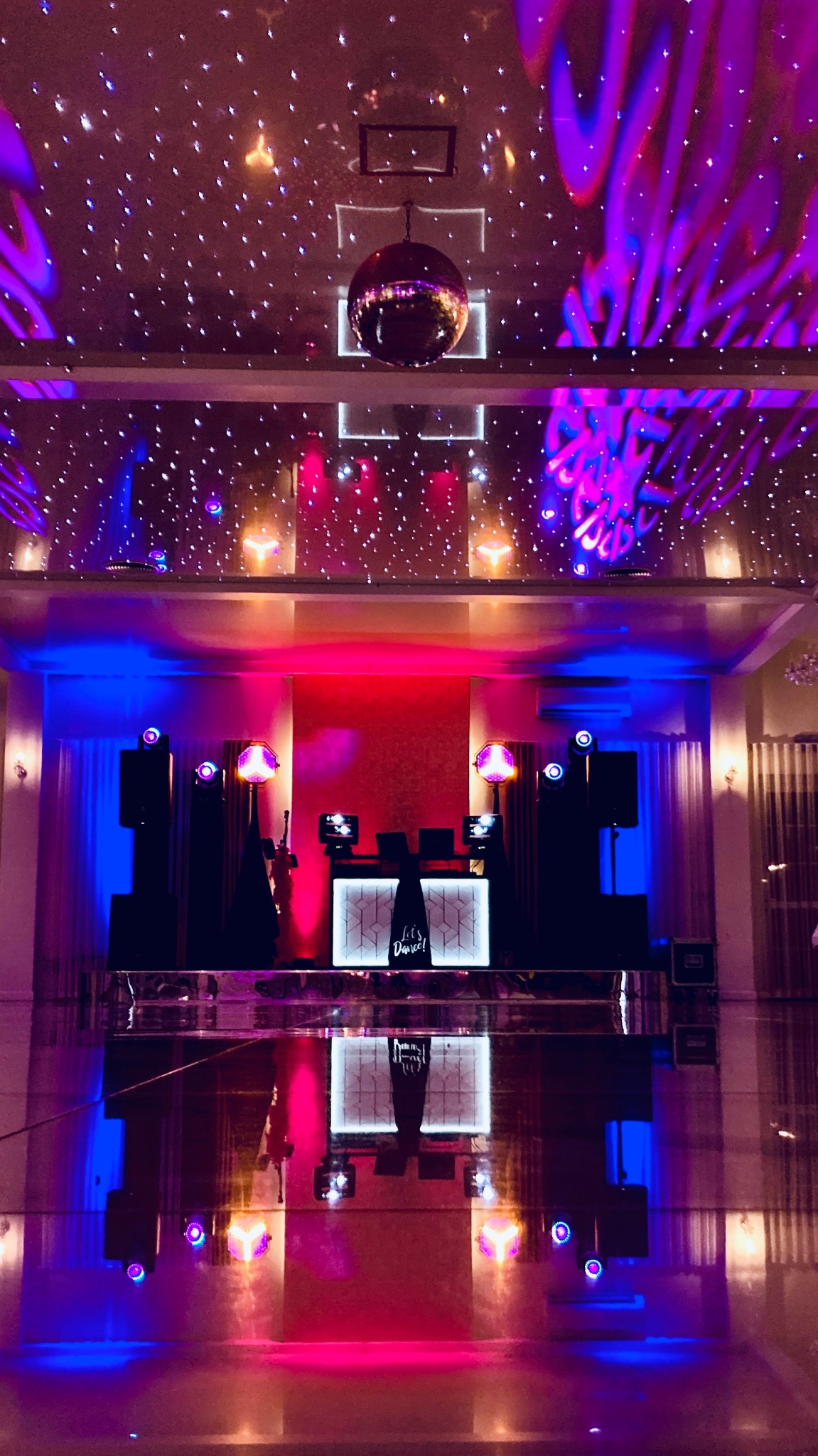 Scena z nagłośnieniem i oświetleniem w fioletowo-niebieskich barwach, lustrzana kula pod sufitem, napis 'Let's Dance!' na podeście DJ-a.