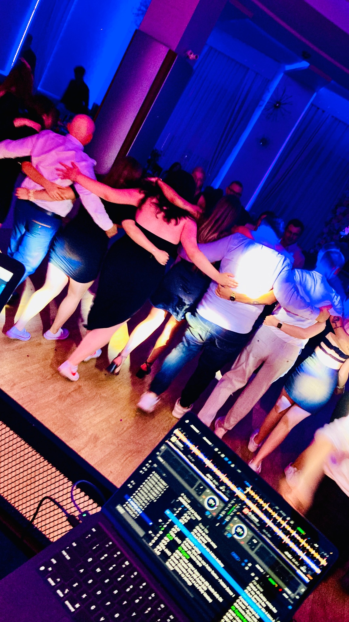 Tańczący w kręgu ludzie, oświetleni niebieskim światłem, z widocznym laptopem DJ-a w rogu kadru. Parkiet, impreza, zabawa.