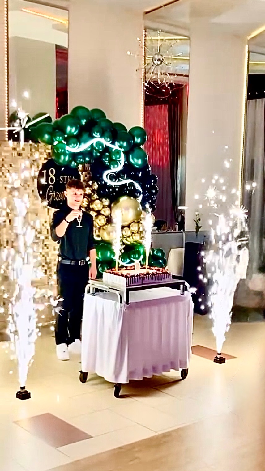 Uroczystość osiemnastych urodzin z tortem, balonami w odcieniach zieleni i złota oraz fontannami iskier na tle dekoracji sali.