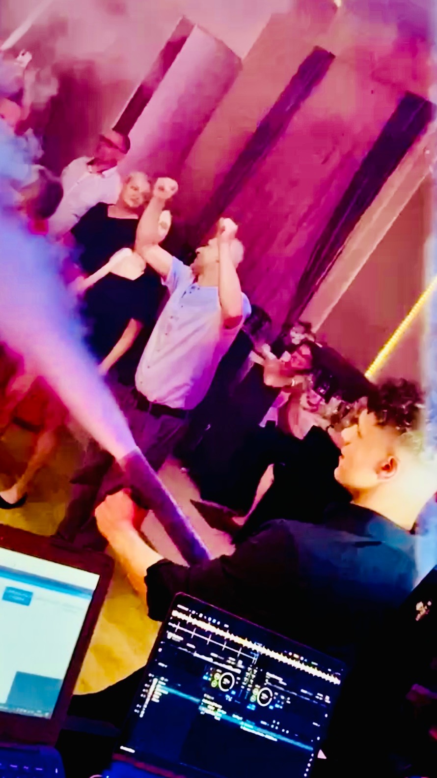 DJ z laptopem i wytwornicą dymu w akcji, rozbawiony tłum w tle, dynamiczne oświetlenie i energia imprezy.