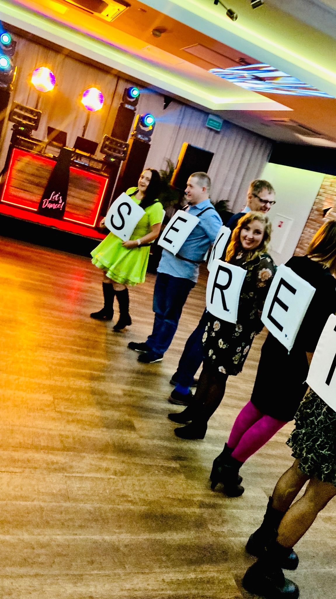 Zespół osób trzymających litery układające się w słowo 'SERDECZNIE' na sali tanecznej z oświetleniem scenicznym. Nietypowe ujęcie z parkietem w roli głównej.