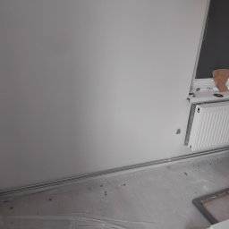 Hydraulik Gazownik - Biały grzejnik panelowy zamontowany pod oknem na tle białej ściany w trakcie remontu. Widoczne rury grzewcze i folia ochronna na podłodze.