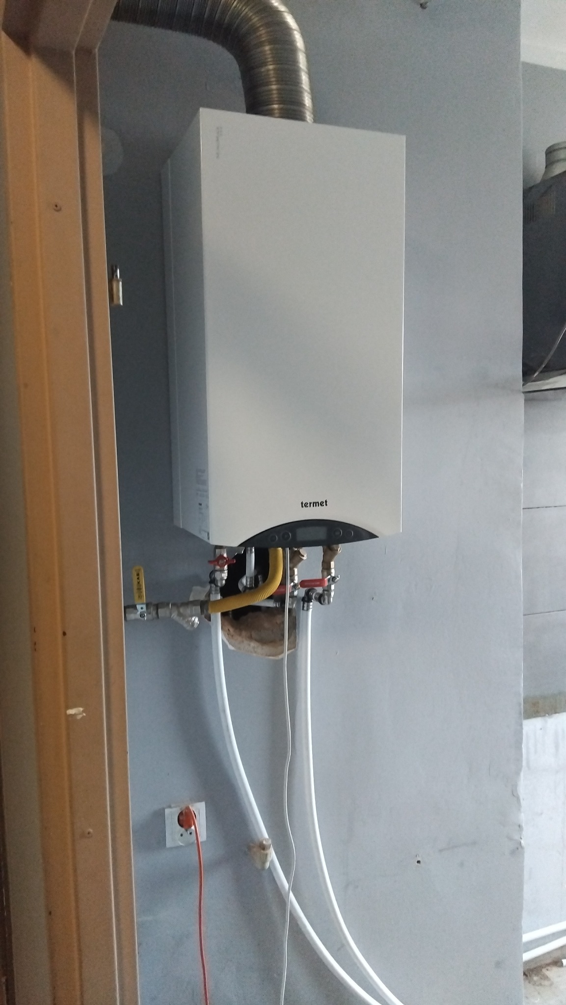 Biały, wiszący piec gazowy marki Termet, z podłączonymi rurami i przewodami, zamontowany na szarej ścianie w pomieszczeniu w trakcie instalacji. Widoczny fragment drzwi i gniazdko.